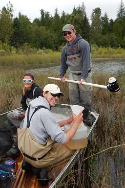 Three students display tools to measure water quality while sitting in a rowboat in a wetland.